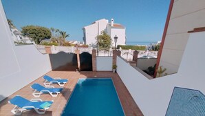 Una piscina en la azotea con sillones azules, vista al mar y un edificio blanco al fondo.