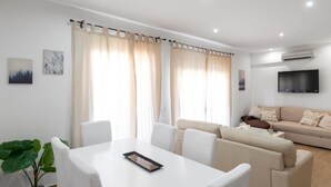 A modern living room with a white sofa, a dining table with chairs, and a large window with curtains.