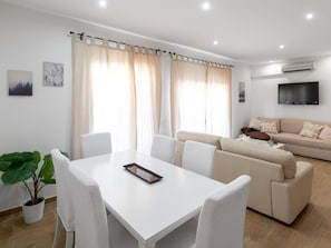 A modern living room with a white sofa, a dining table with chairs, and a large window with curtains.