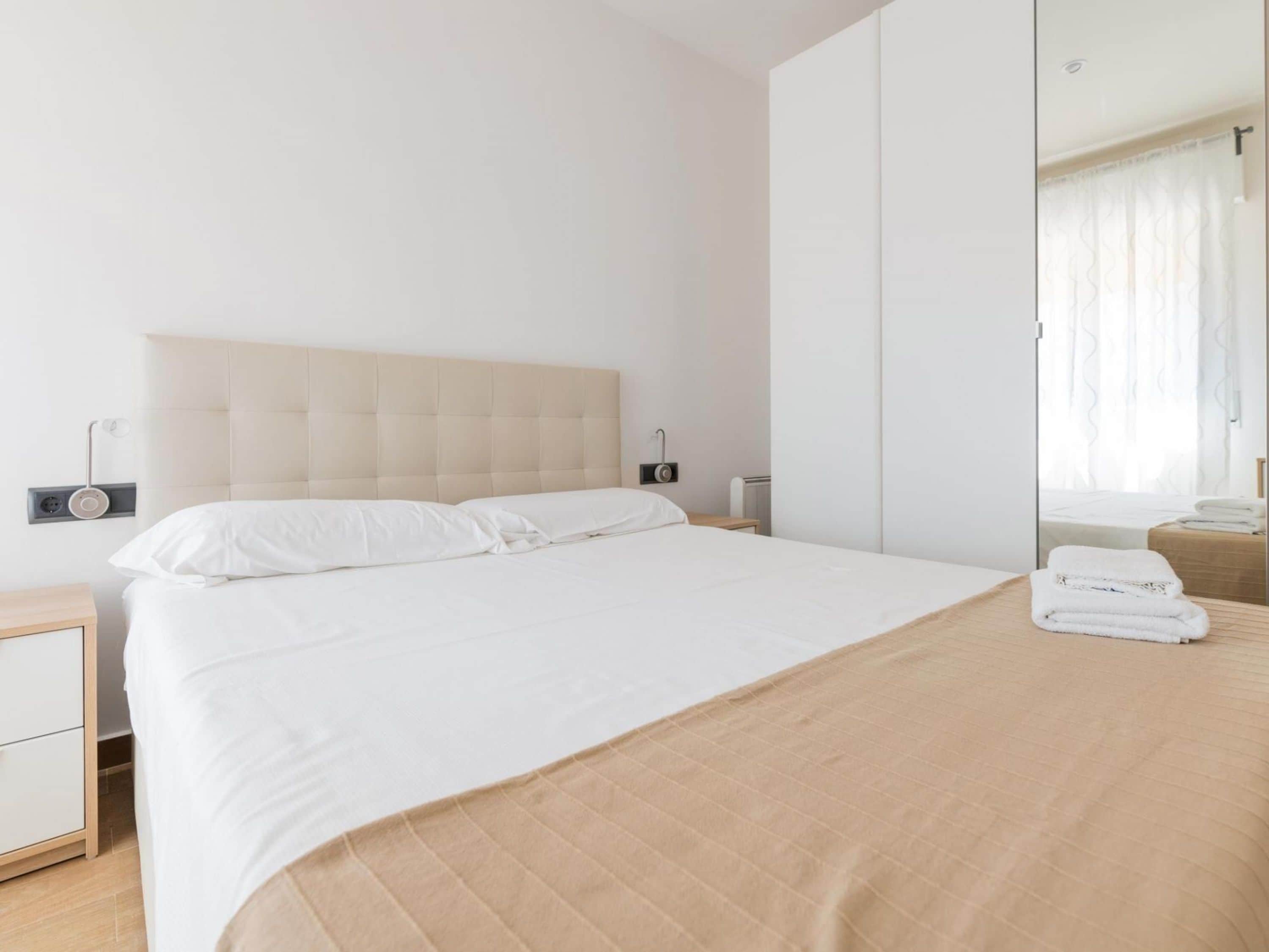 Una cama bien hecha con sábanas blancas y cabecera color beige, una mesita de noche con lámpara, y un espejo que refleja una ventana con cortinas.