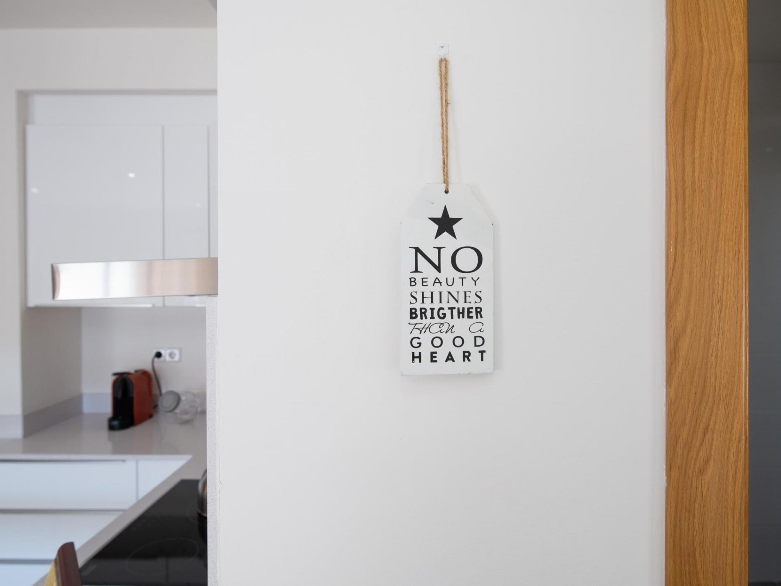 Une cuisine avec des meubles blancs, un panneau décoratif suspendu portant une citation inspirante et un cadre de porte en bois.