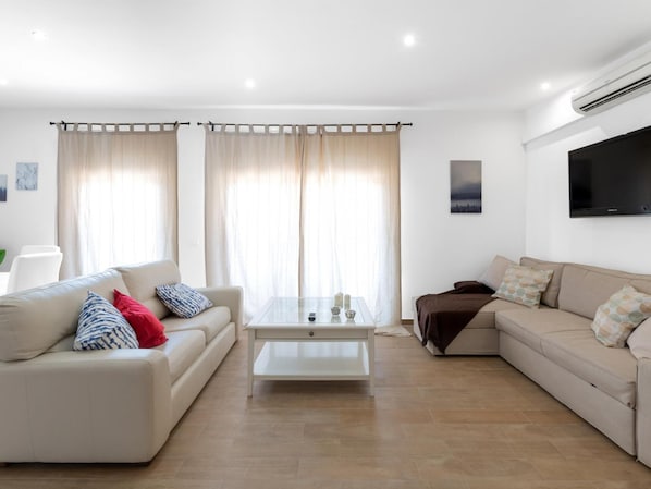 A modern living room with a beige sofa, a white coffee table, and a brown sofa. There is a flat-screen TV mounted on the wall.