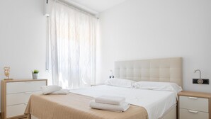 A bedroom with a large bed, a bedside table, a nightstand, a lamp, and a window with curtains. - Maka Beach Apartment V (Castelldefels)