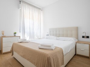 A bedroom with a large bed, a bedside table, a nightstand, a lamp, and a window with curtains. - Maka Beach Apartment V (Castelldefels)