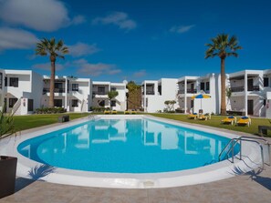 A swimming pool surrounded by white buildings with balconies, palm trees, and sun loungers.