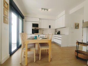 Una cocina moderna con armarios blancos, una zona de comedor con mesa y sillas de madera, y un ventanal que proporciona luz natural.