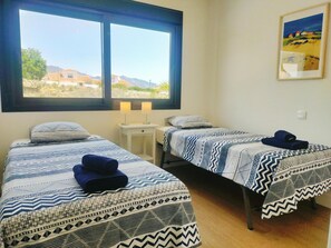 A room with two beds, each with patterned bedding, a window with a view of buildings and hills, and a painting on the wall.