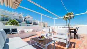 Una terraza en la azotea con mobiliario exterior blanco, una mesa de madera con fruta y vistas a un edificio blanco y palmeras.