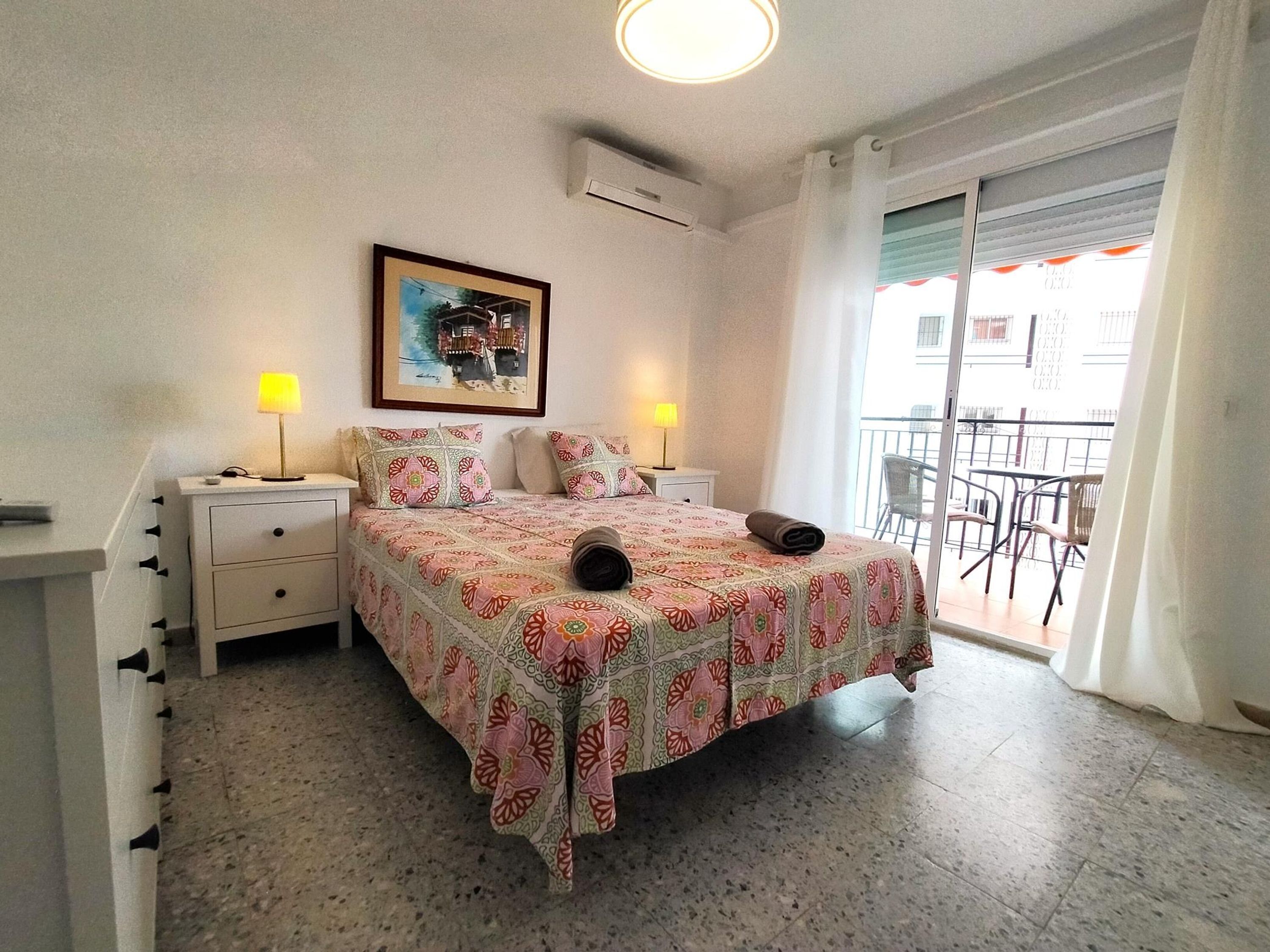 Un dormitorio con cama, dos mesitas de noche con lámparas, un cuadro enmarcado en la pared y un balcón con sillas.