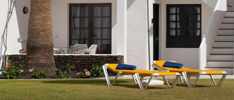 A poolside area with two sun loungers under a blue and yellow umbrella, a palm tree, and a white building with a stone wall and a window with curtains.