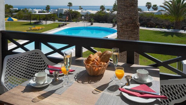 A dining table with breakfast items, a view of a pool, and a palm tree. - Apt 32 Las Malvas (Haria)