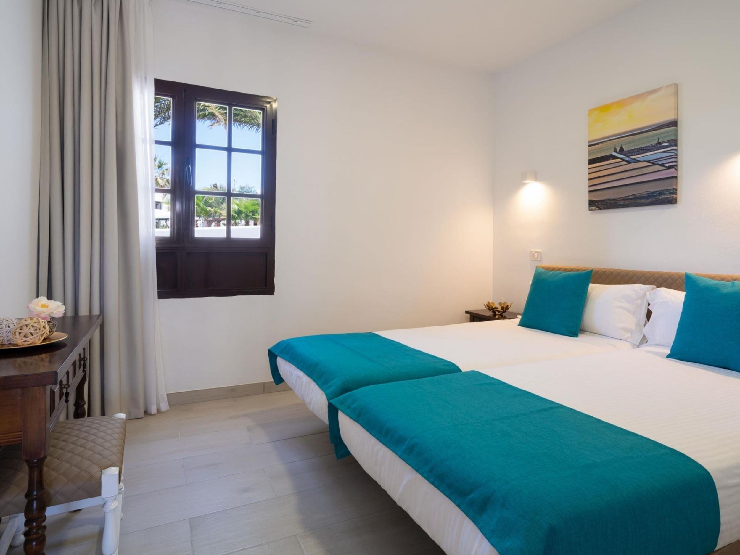 A hotel room with a large bed, a desk with a chair, a window with curtains, and a painting on the wall.