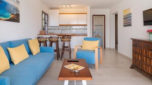 A modern living room with a blue sofa, wooden chairs, a coffee table, and a kitchen in the background. - Apt 32 Las Malvas (Haria)