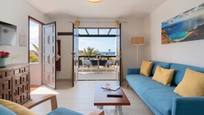 A modern living room with a blue sofa, a wooden coffee table, and a view of the sea through an open door. - Apt 32 Las Malvas (Haria)