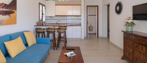 Uma sala de estar moderna com um sofá azul, mesa de centro de madeira e uma cozinha ao fundo.