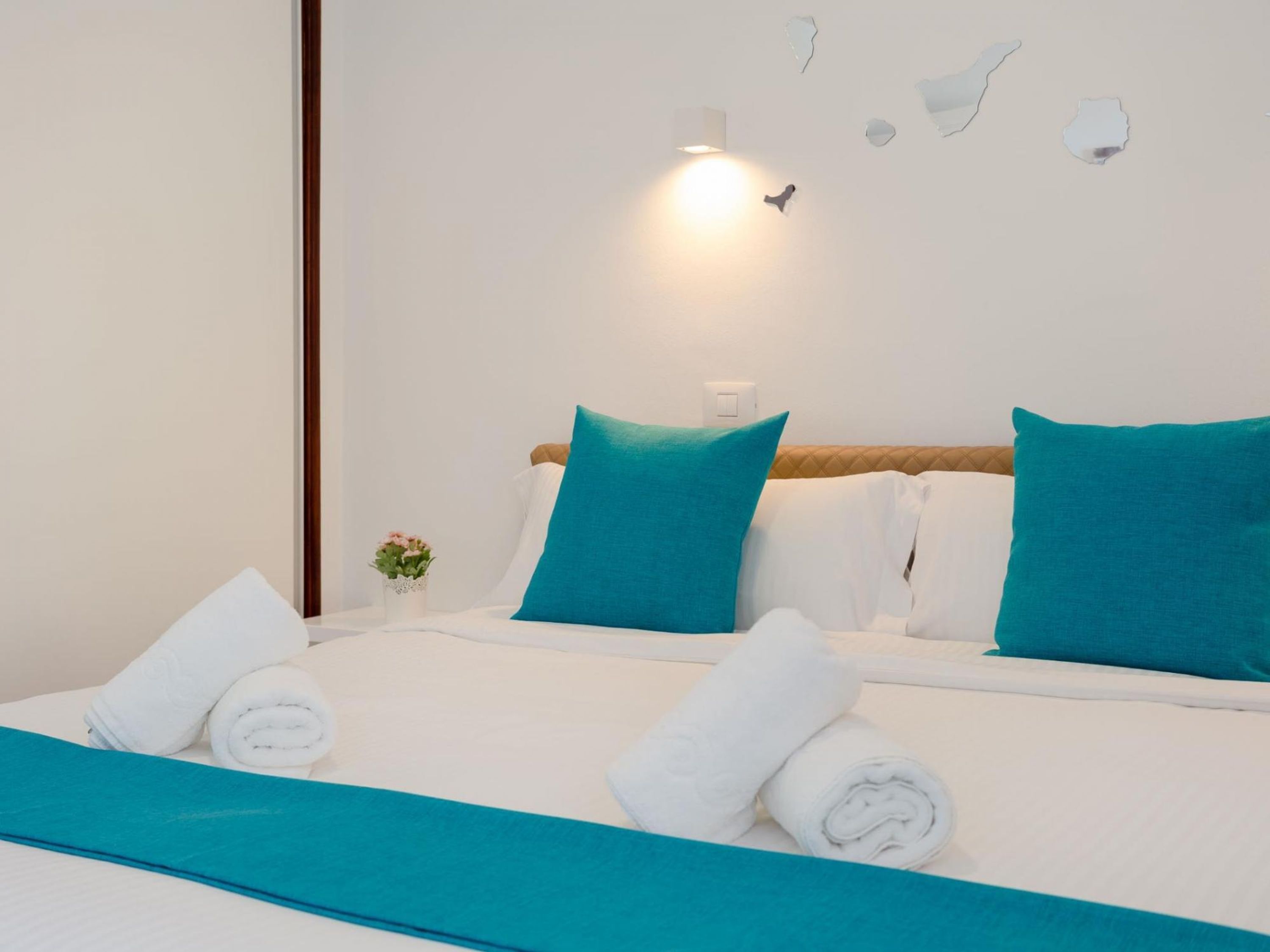 A neatly made bed with white linens and teal pillows, a small vase with flowers, and a wall-mounted light fixture.