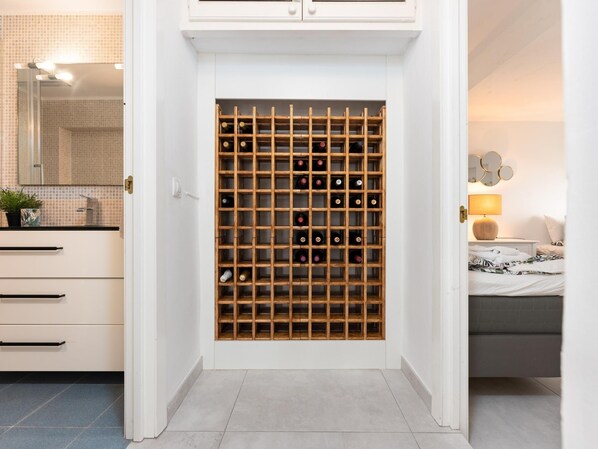 A hotel room with a wine rack, a bed, a bedside table with a lamp, and a bathroom with a mirror and sink.