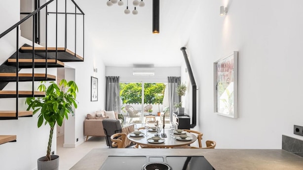 A modern kitchen with a central island, a staircase with black railings, and a dining area with a round table and chairs. - Agrupadas Beautiful Duplex With Amazing Location (Marbella)