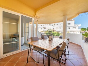 Ein Balkon mit Holztisch und -stühlen, eine Glastür, die zu einem Raum führt, und im Hintergrund ein Blick auf Gebäude.