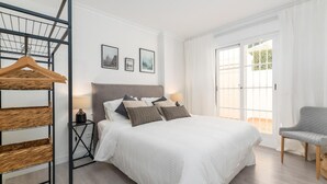 A bedroom with a bed, a chair, a hanging rack, and a window with curtains. - Se or o de Aloha 2 Bedroom (Marbella)