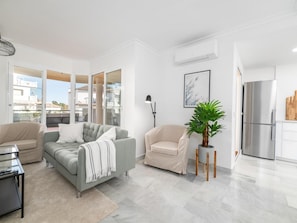 A modern living room with a grey sofa, a beige armchair, a small potted plant, and a kitchen area with a stainless steel refrigerator. - Se or o de Aloha 2 Bedroom (Marbella)
