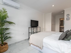 A modern bedroom with a bed, a television on a stand, a potted plant, and a bathroom visible through an open door.