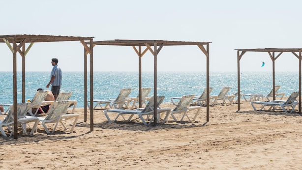 A beach with sun loungers under wooden structures, a person standing, and a clear view of the sea. - Beachfront Sunny Apartment Sea Mountain View (Castelldefels)