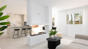 A modern kitchen with white cabinetry, a marble backsplash, and a central island with bar stools. There is a living area with a sofa, a coffee table, and a potted plant.
