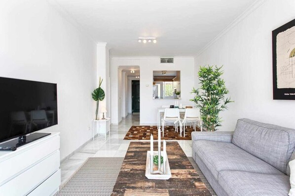 A modern living room with a flat-screen TV, a grey sofa, a coffee table, and a dining area in the background. - Senorio de Gonzaga Great 2 Bedroom Apartment (Marbella)
