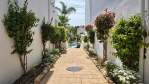 Un estrecho sendero bordeado por paredes blancas y diversas plantas, que conduce a una zona de piscina.