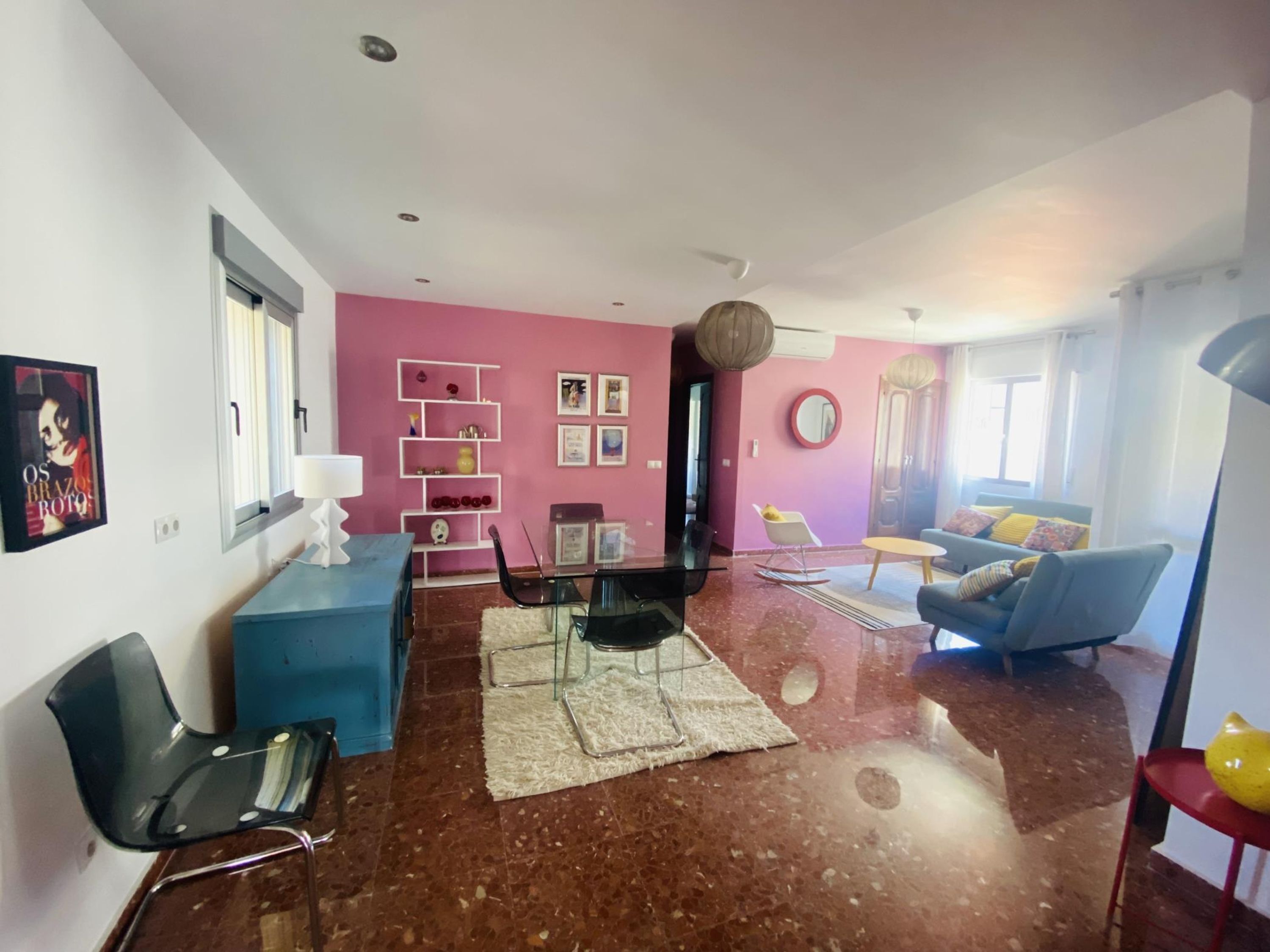 Ein modernes Wohnzimmer mit einer rosa Wand, einem blauen Sofa, einem Essbereich mit einem Glastisch und einer Küchenzeile mit einem weißen Schrank.