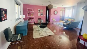 A modern living room with a pink wall, a blue sofa, a dining area with a glass table, and a kitchen area with a white cabinet. - Nerja Sun Apartamento Jabega Torrecilla (Nerja)