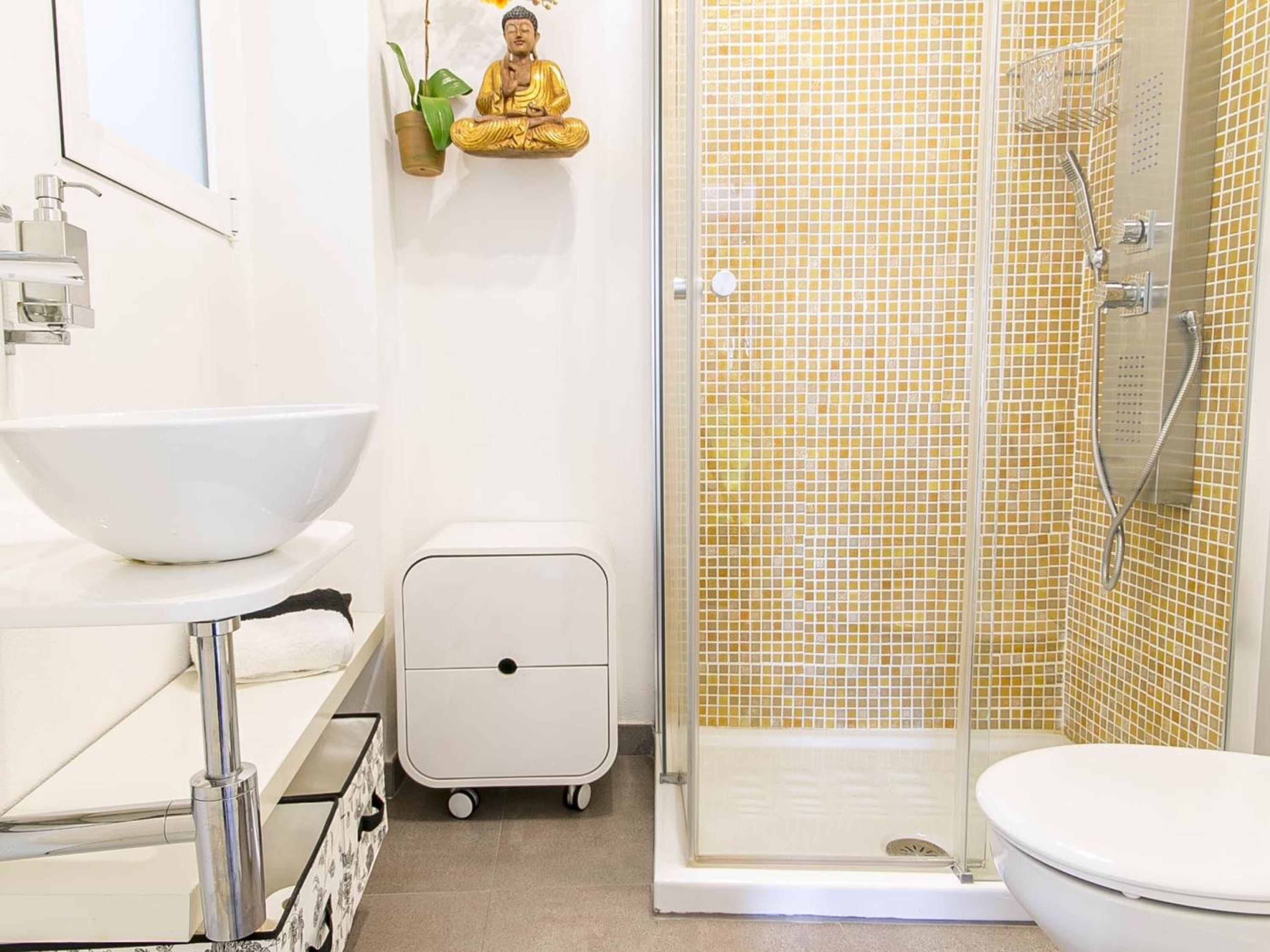 Ein Badezimmer mit einem weißen Waschbecken, einer Toilette, einer Dusche mit gelben Mosaikfliesen und einer kleinen Buddha-Statue.