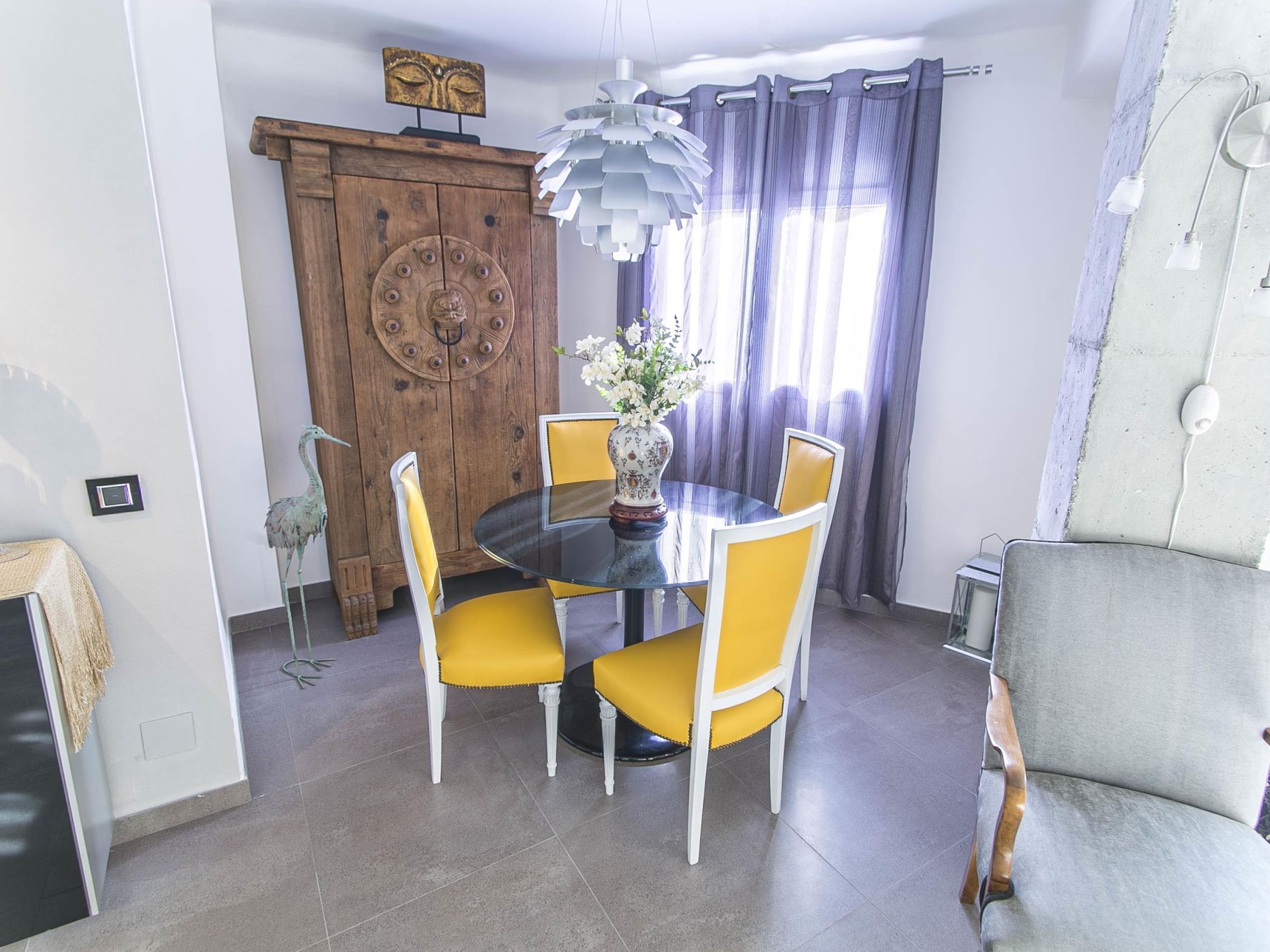 A dining area with a round glass table, yellow chairs, a wooden cabinet with a decorative door, and a hanging light fixture.