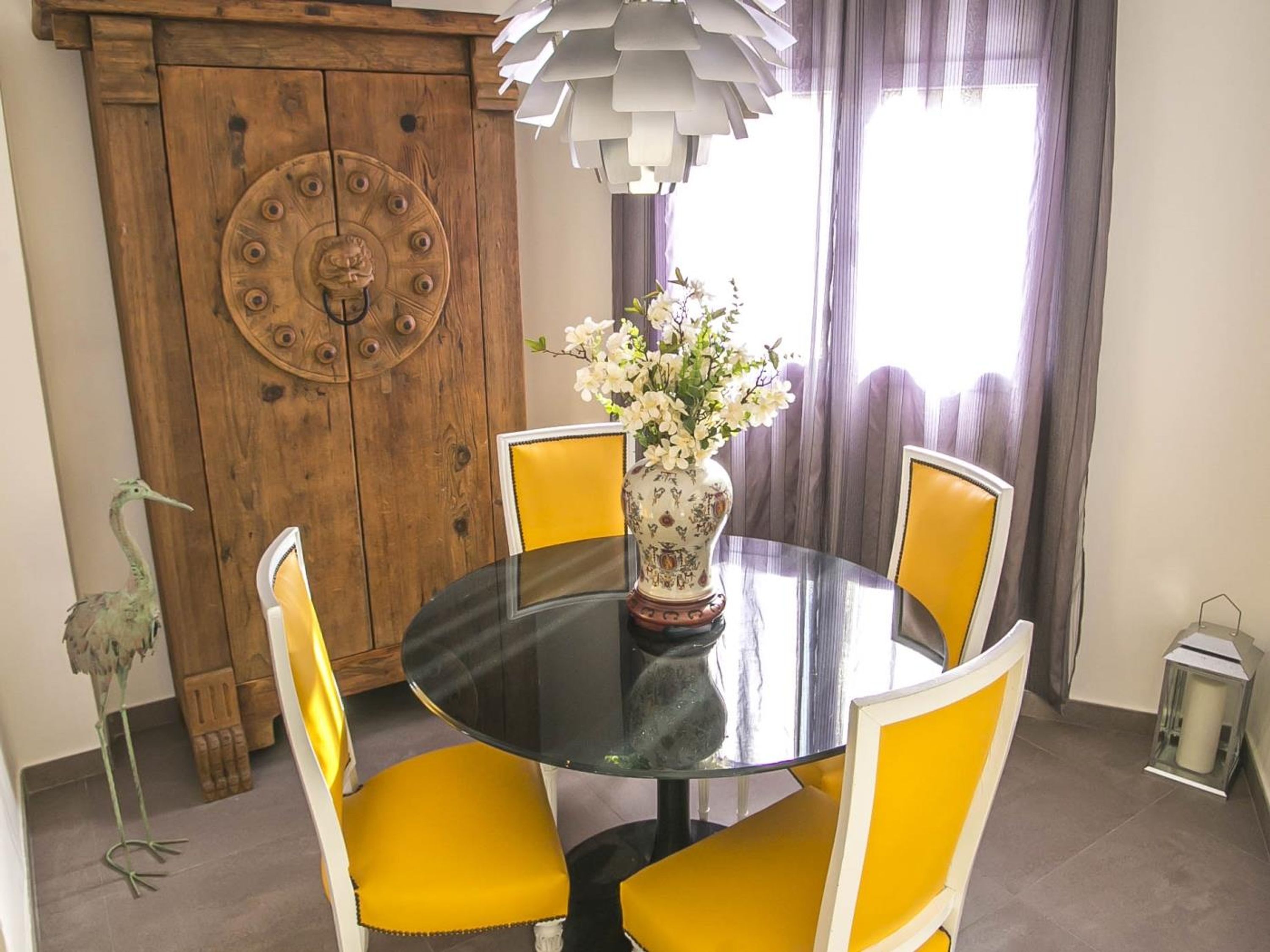 A dining area with a round table, yellow chairs, a vase with flowers, a wooden door, and a hanging light fixture.