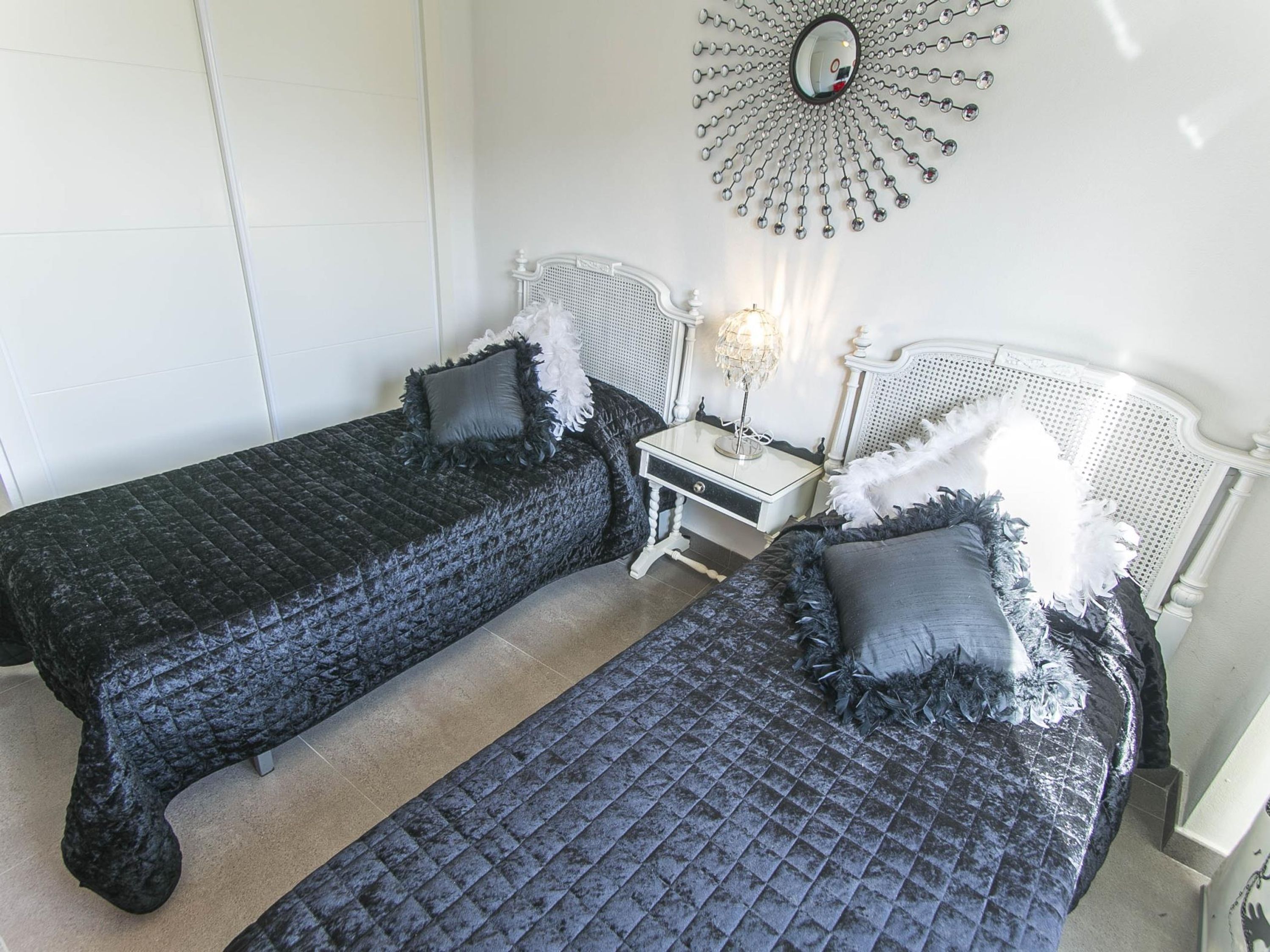 Une chambre avec deux lits, chacun recouvert d’une courtepointe matelassée de couleur sombre et agrémenté d’oreillers assortis. Une table de chevet blanche avec une lampe se trouve entre les deux lits.