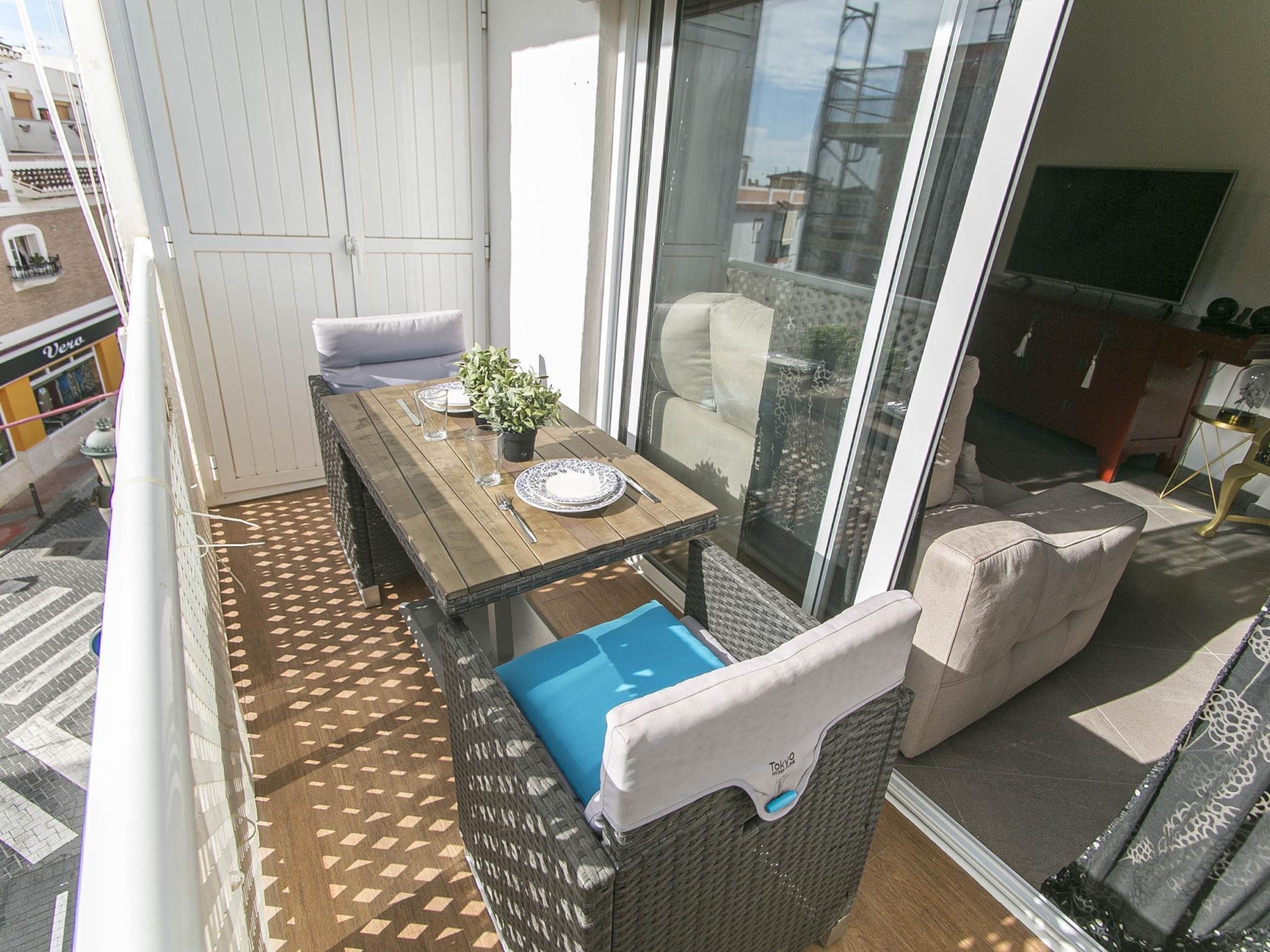 A balcony with a wooden table set for two, a wicker sofa, and a view of the city through large glass doors.