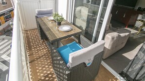 A balcony with a wooden table set for two, a wicker sofa, and a view of the city through large glass doors.