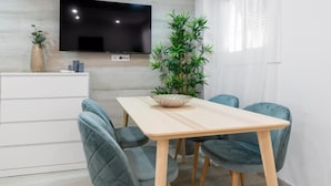 Een moderne eetruimte met een houten tafel, vier stoelen, een flatscreen televisie aan de muur, een potplant en een witte kast.