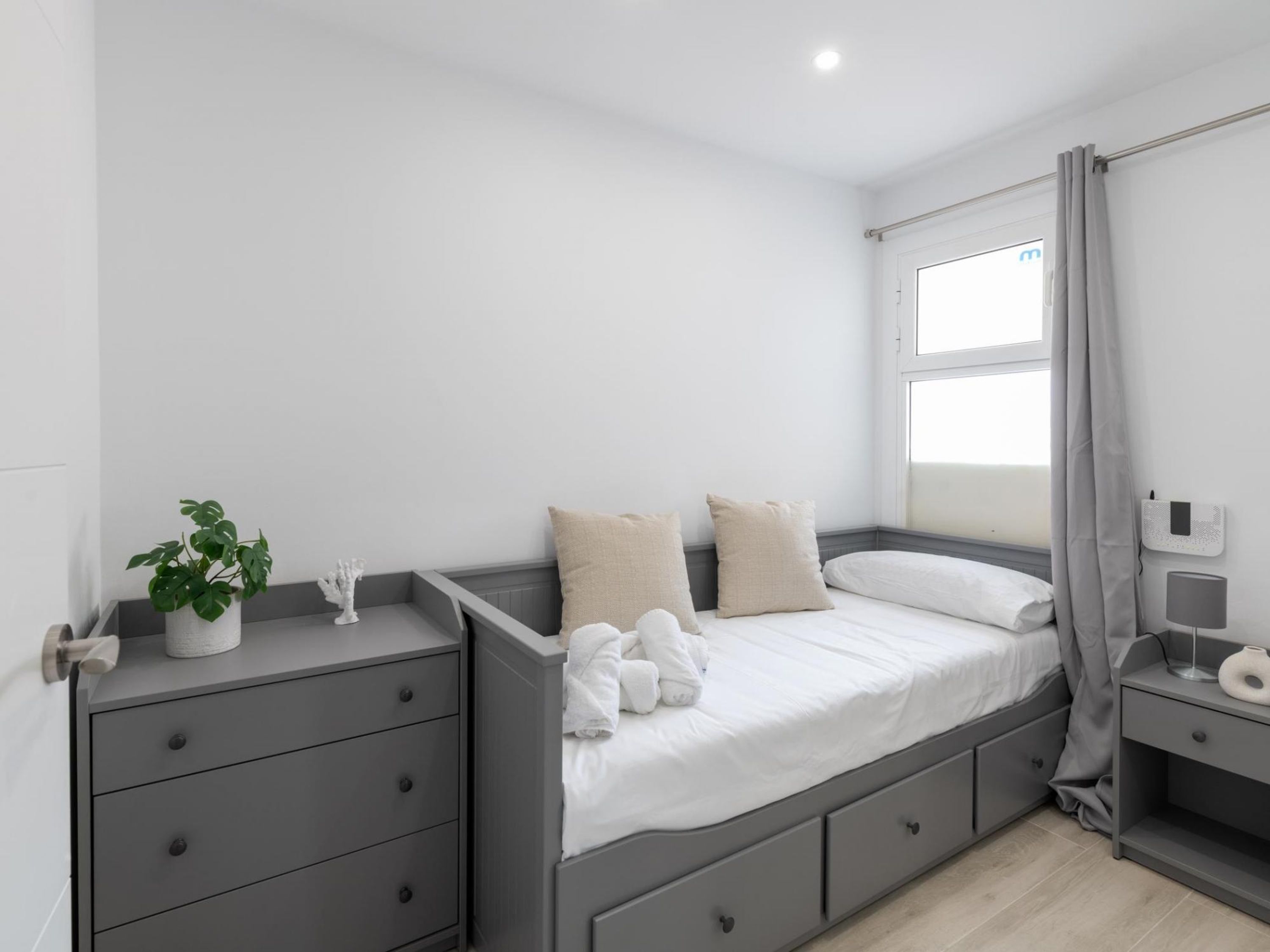 A modern bedroom with a grey bed, white bedding, a grey nightstand, a bedside table, a potted plant, and a window with grey curtains.