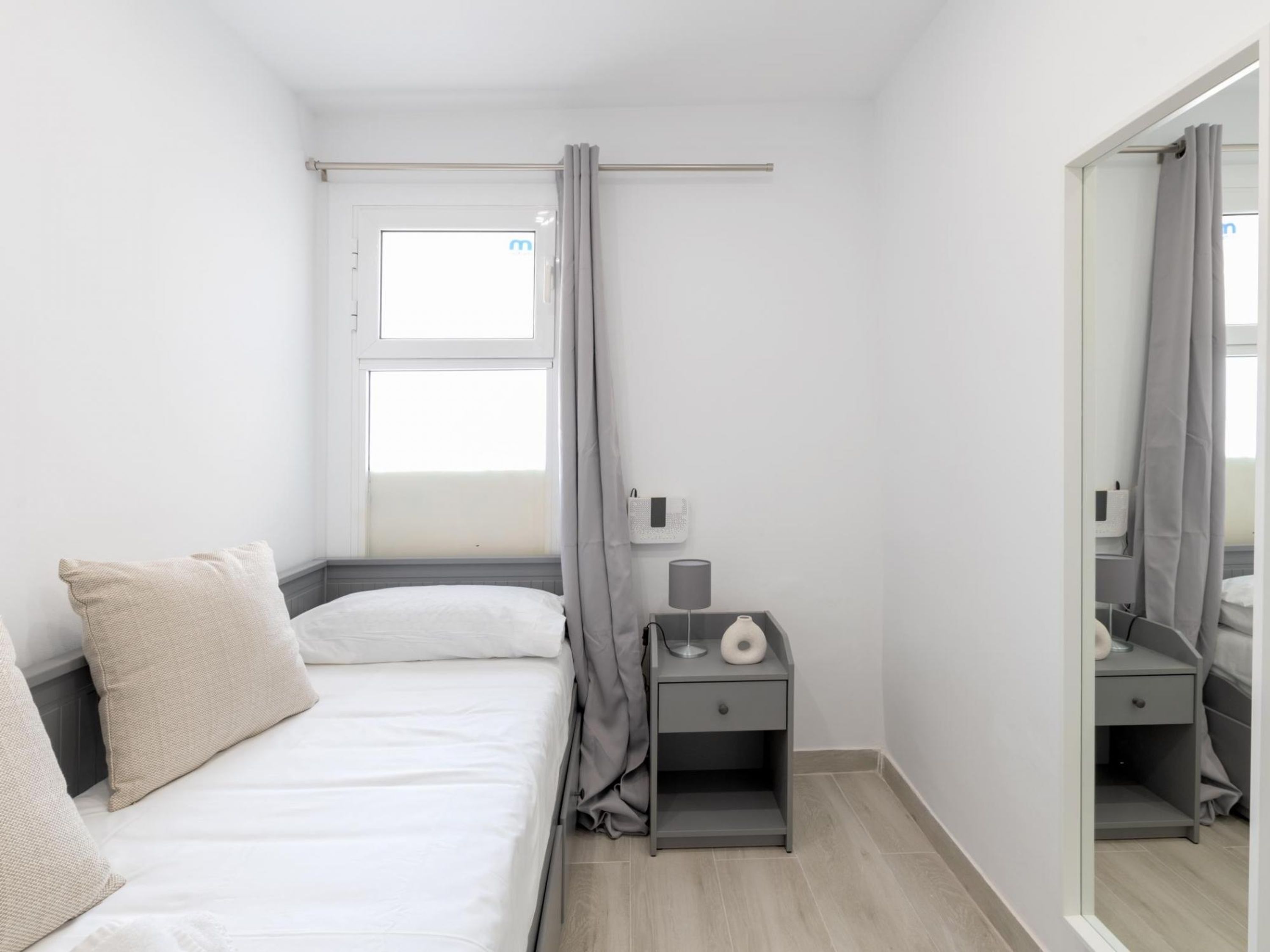 Un dormitorio pequeño y moderno con cama, mesita de noche, espejo y ventana con cortinas.