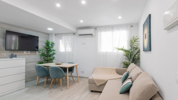 Een moderne woonkamer met een beige bank, een eethoek met een tafel en stoelen, een flatscreen televisie aan de wand en een groot raam met gordijnen.