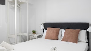 Een net opgemaakt bed met witte en bruine kussens, een klein nachtkastje met een plantje, en een witte kledingkast.