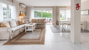 A spacious living room with a beige sofa, a coffee table, and a patterned rug. There is a dining area with white chairs and a table, and a red decorative item on the wall. - Sol y Paz 3 Family Apartment With Great Location (Marbella)