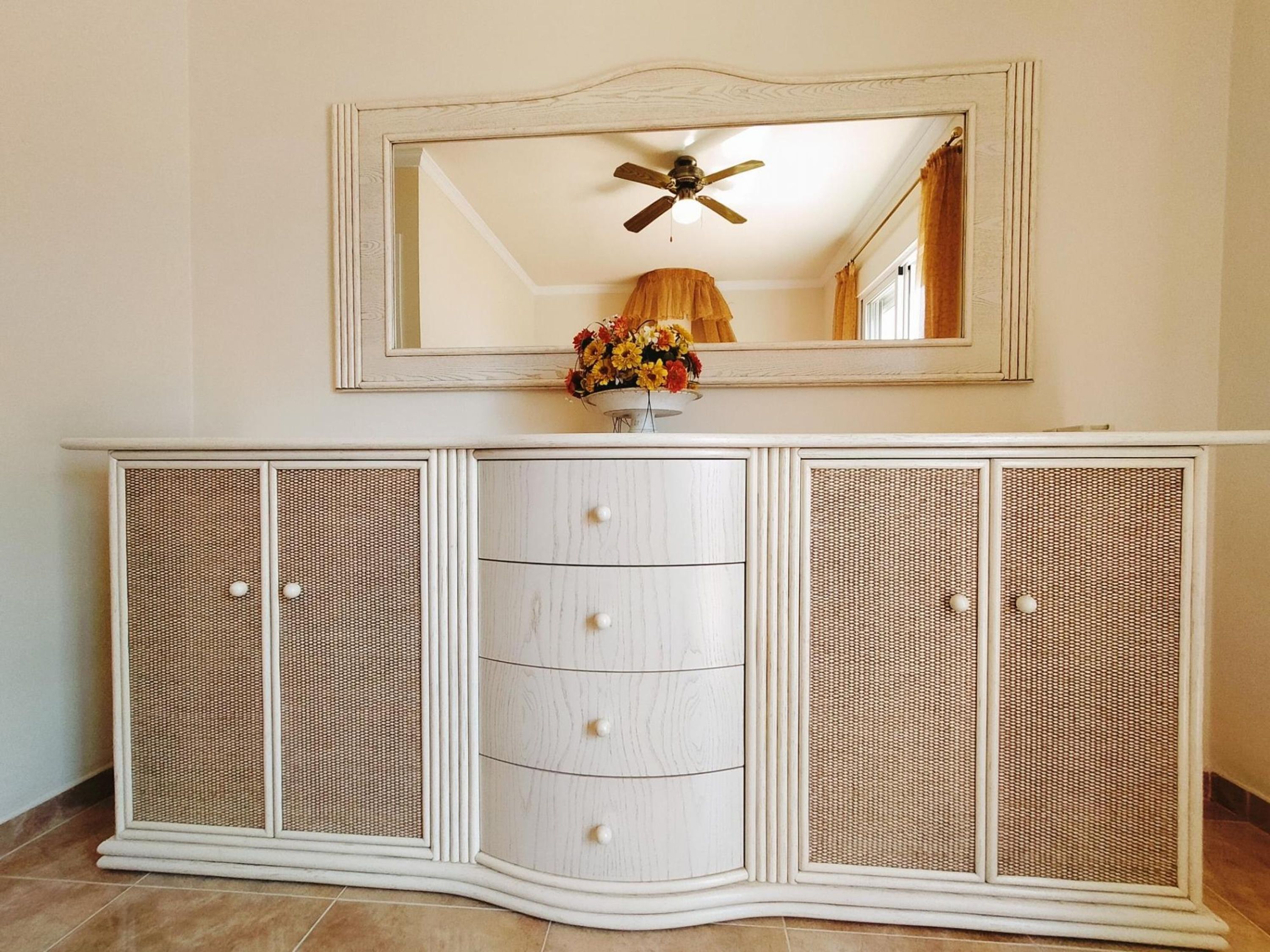 A white cabinet with a mirror above it, a ceiling fan, and a vase with flowers.