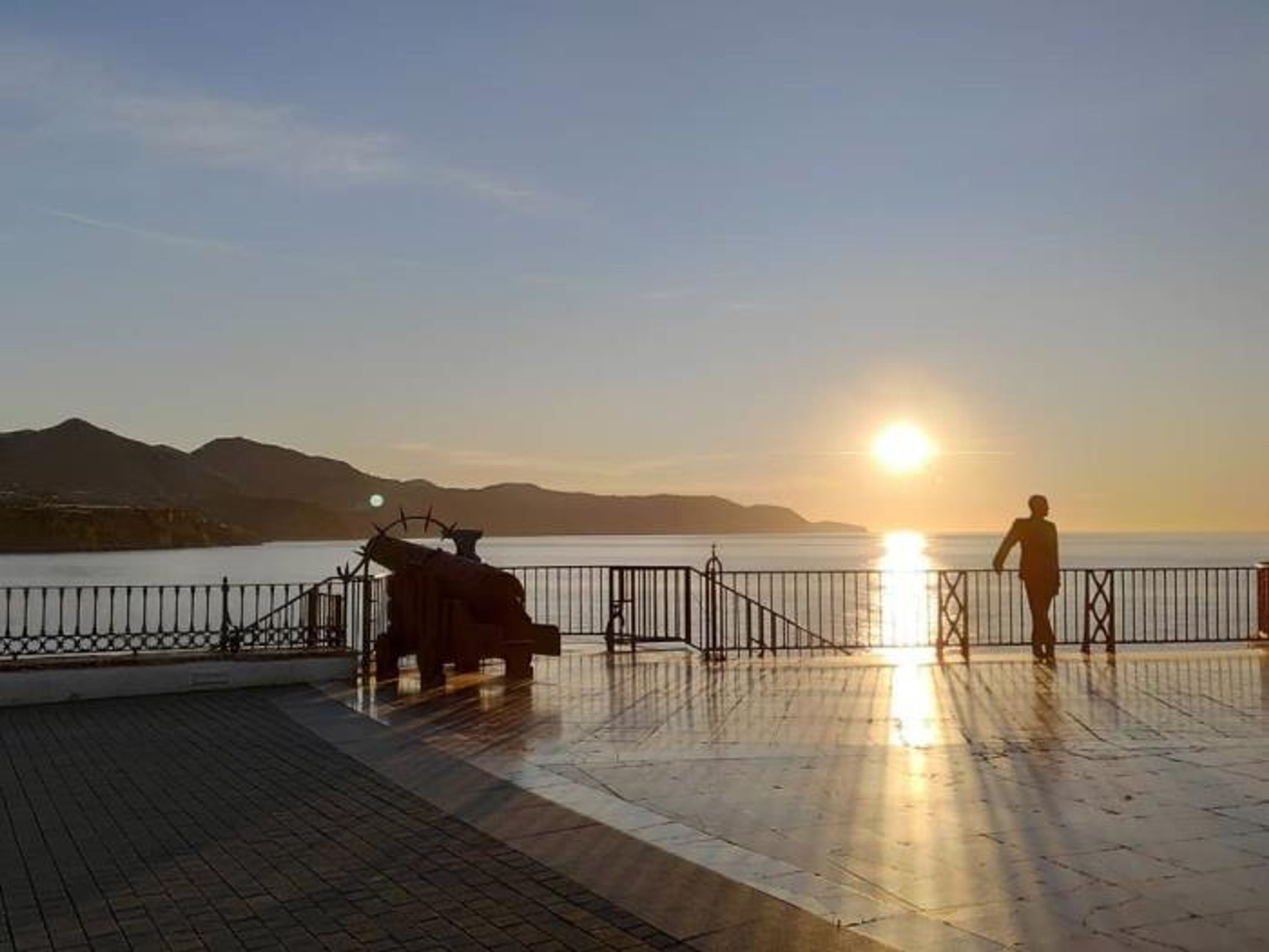 A waterfront promenade with a metal fence, a bench, and a person standing facing the sunset.
