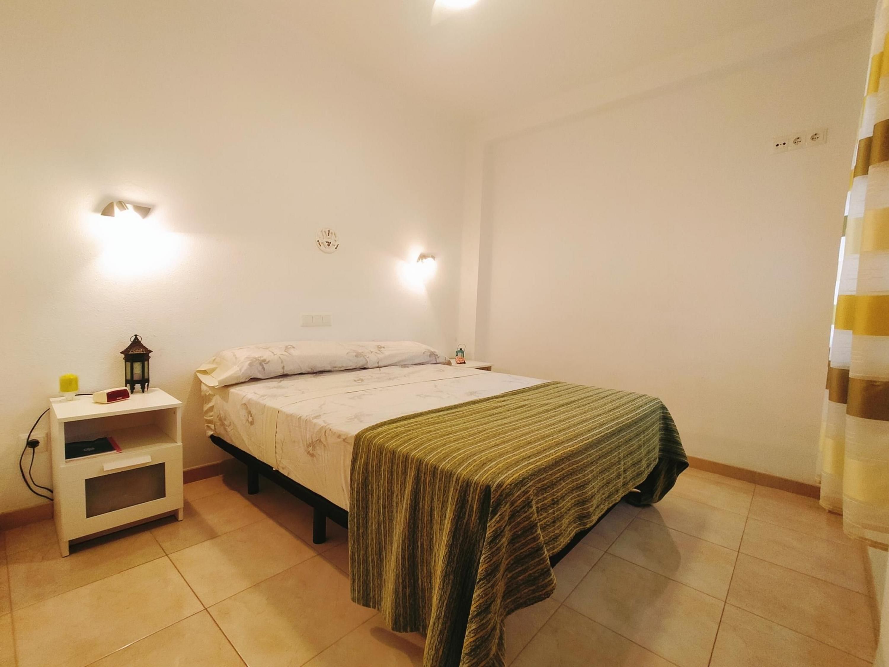 A bedroom with a bed, bedside table, lamp, and wall-mounted light.