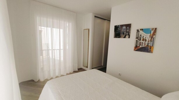 A hotel room with a bed, a large window with sheer curtains, a door, and two wall-mounted pictures. - Apartamento Bronce B Centro Nerja Sun (Nerja)