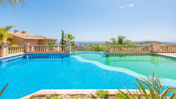 A clear blue swimming pool with a curved edge, surrounded by a stone balustrade, set against a backdrop of a clear sky and distant landscape.