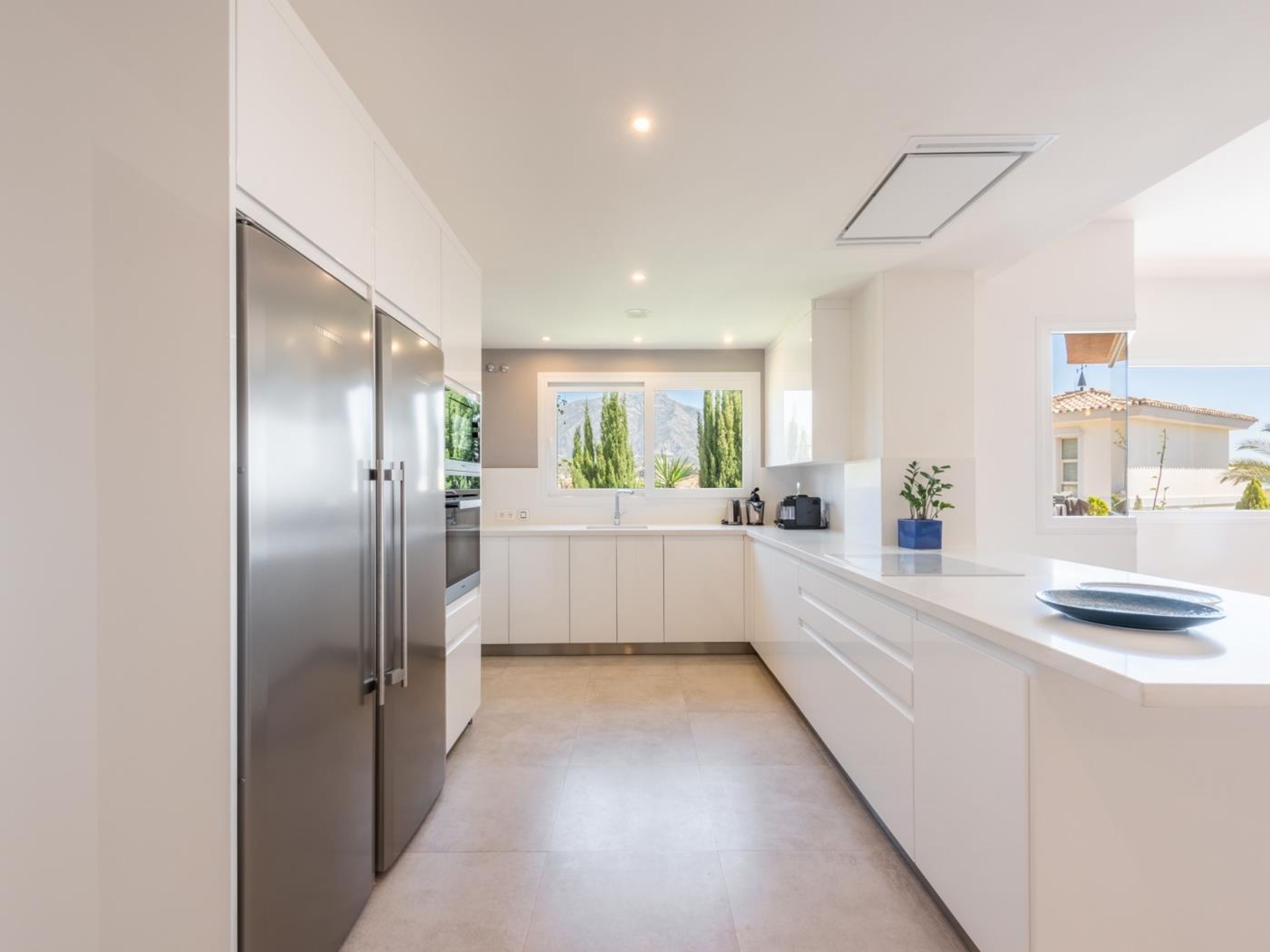 Cocina moderna con muebles blancos, refrigerador de acero inoxidable y ventana con vistas a zonas verdes y montañas.